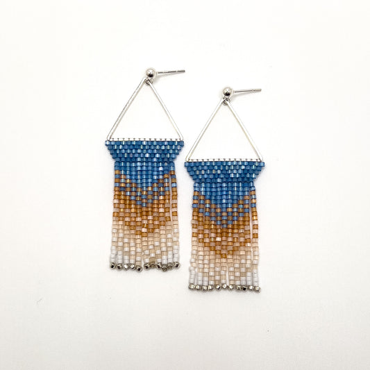 Pair of beaded Eastern Bluebird earrings laying on a white background, featuring a triangular silver frame with a solid blue beaded top that transitions into amber and white beaded fringe, creating a gentle gradient effect.