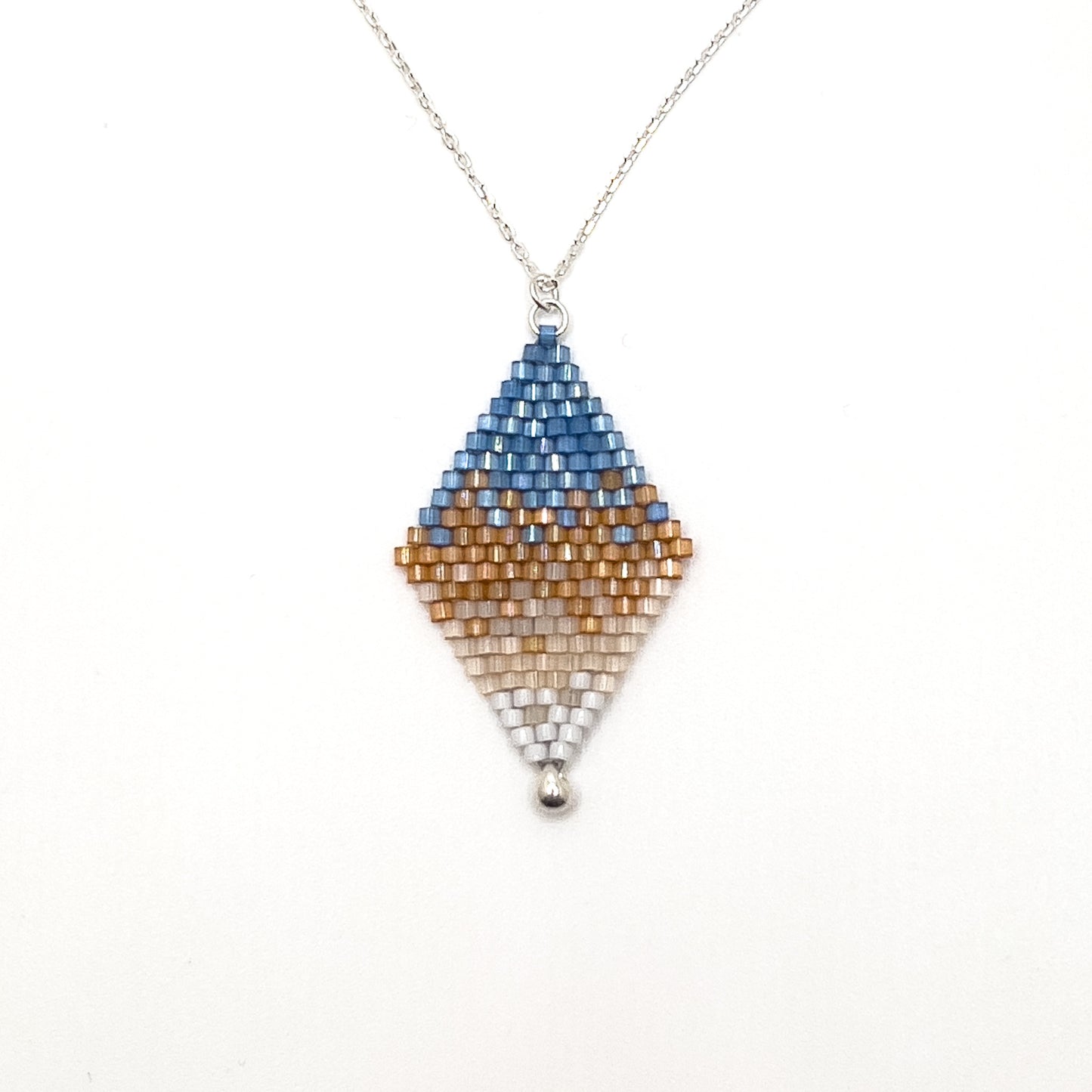 Close up of American Goldfinch-inspired handcrafted beaded pendant with a sterling silver chain displayed on white surface, showcasing a gradient pattern with blue, amber, and white beads.