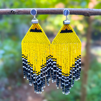 Handcrafted beaded earrings inspired by the American Goldfinch, featuring vibrant yellow, black, and white beads, hanging from a branch against a blurred natural background.