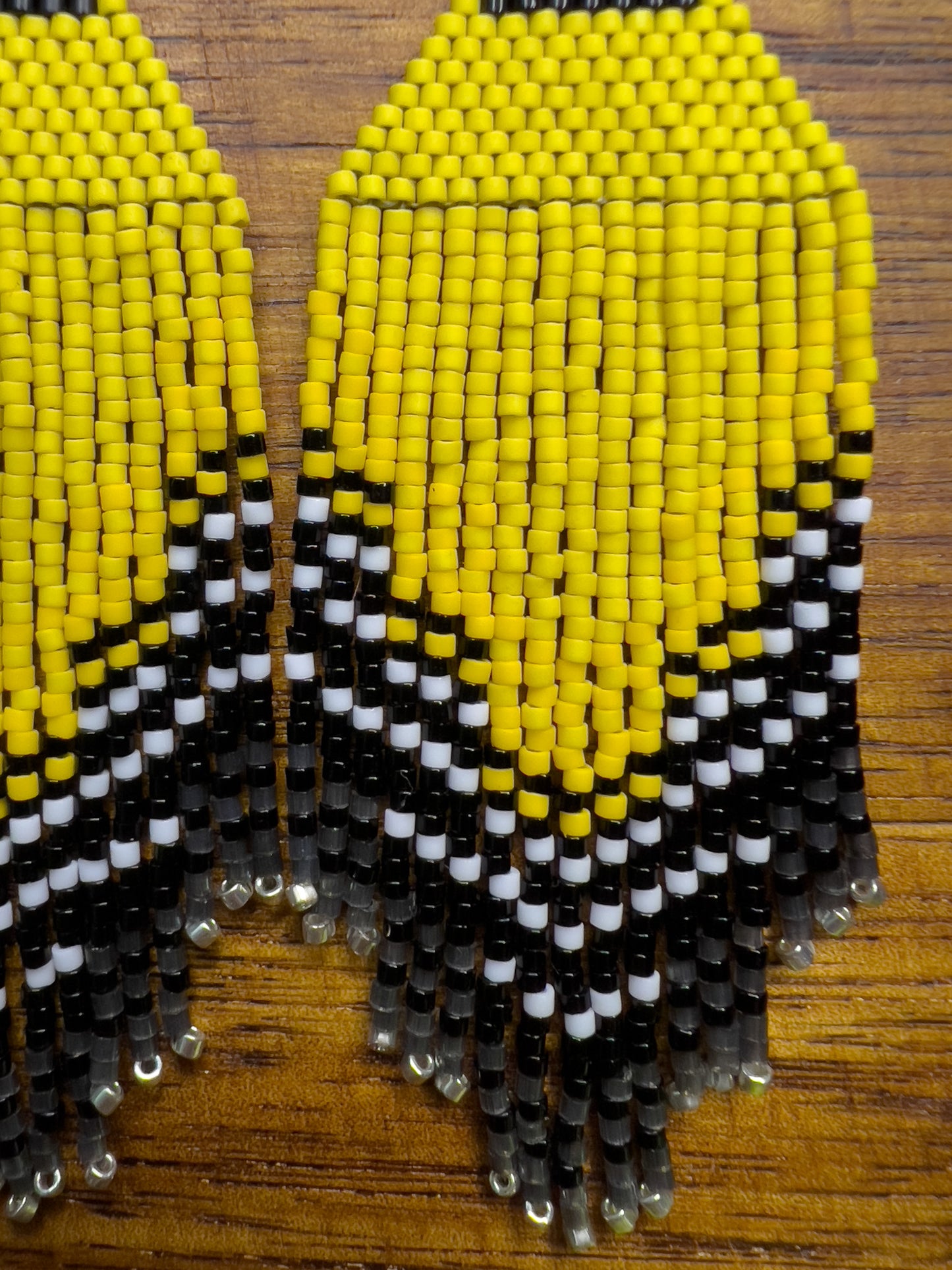 Close up of American Goldfinch-inspired handcrafted beaded earrings displayed on a wooden surface, showcasing bright yellow, black, and white patterns with fringe detail.