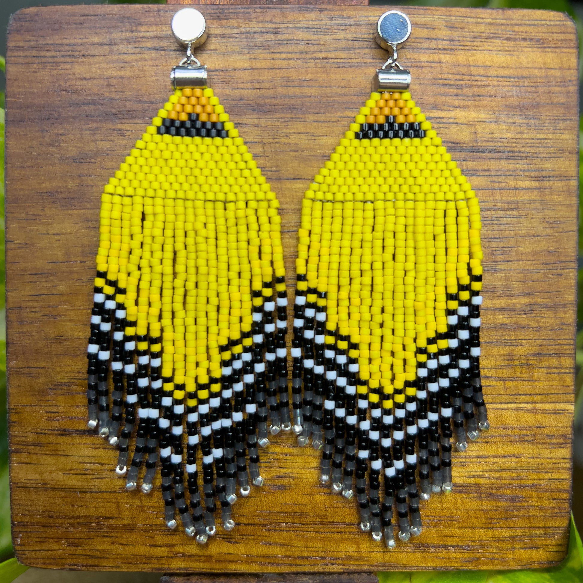 American Goldfinch-inspired handcrafted beaded earrings displayed on a wooden surface, showcasing bright yellow, black, and white patterns with fringe detail.