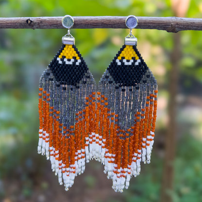 Handcrafted beaded earrings representing an American Robin with a bold triangular design with black, yellow, orange, and white beads, hanging from a branch against a natural, blurred background.