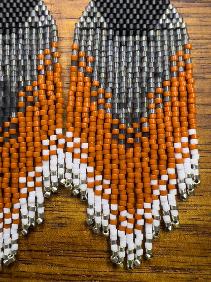 Close up of handcrafted beaded earrings representing an American Robin displayed on a wooden surface, showcasing a vibrant triangular pattern in black, yellow, orange, and white beads with intricate fringe detail.