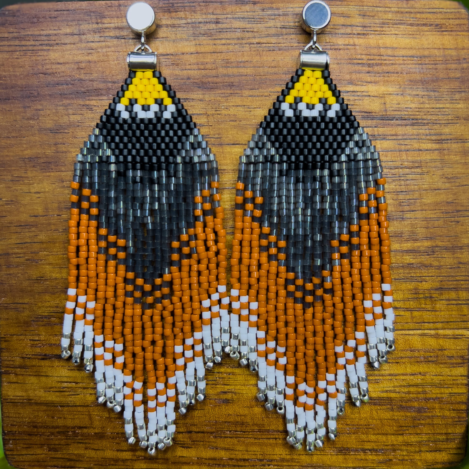 Handcrafted beaded earrings representing an American Robin displayed on a wooden surface, showcasing a vibrant triangular pattern in black, yellow, orange, and white beads with intricate fringe detail.