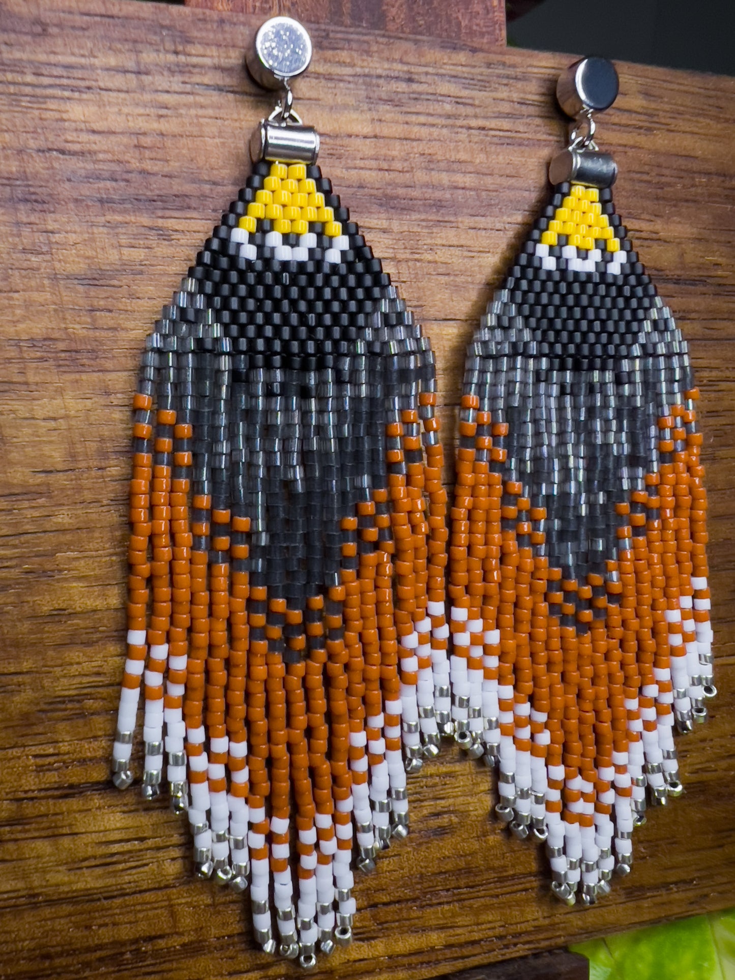 Side view of handcrafted beaded earrings representing an American Robin displayed on a wooden surface, showcasing a vibrant triangular pattern in black, yellow, orange, and white beads with intricate fringe detail.