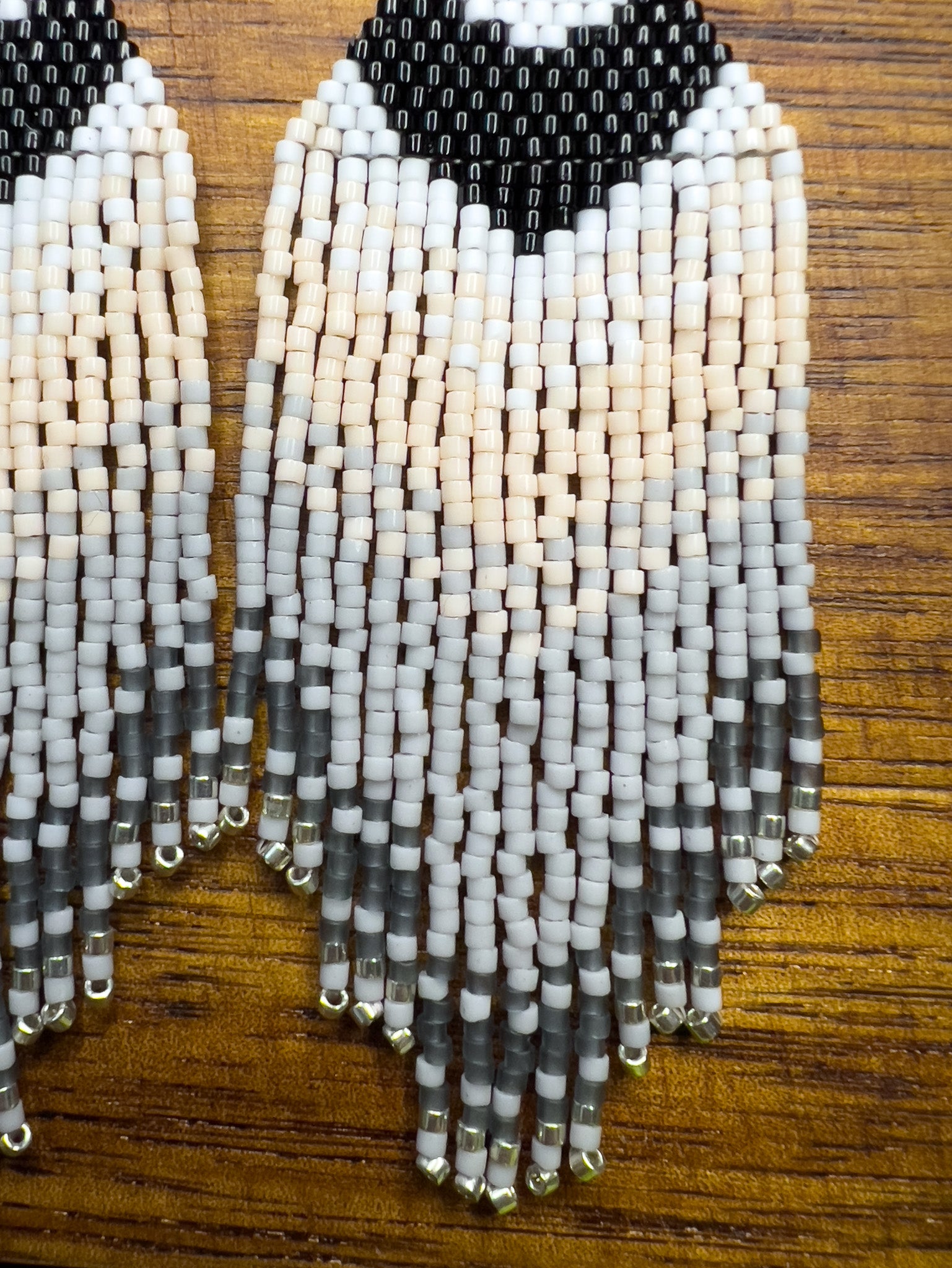 Close up of Carolina Chickadee inspired beaded earrings on a wooden background, featuring a bold black and white V-shape at the top that resembles the bird’s distinctive head pattern, followed by soft cream and gray fringe.