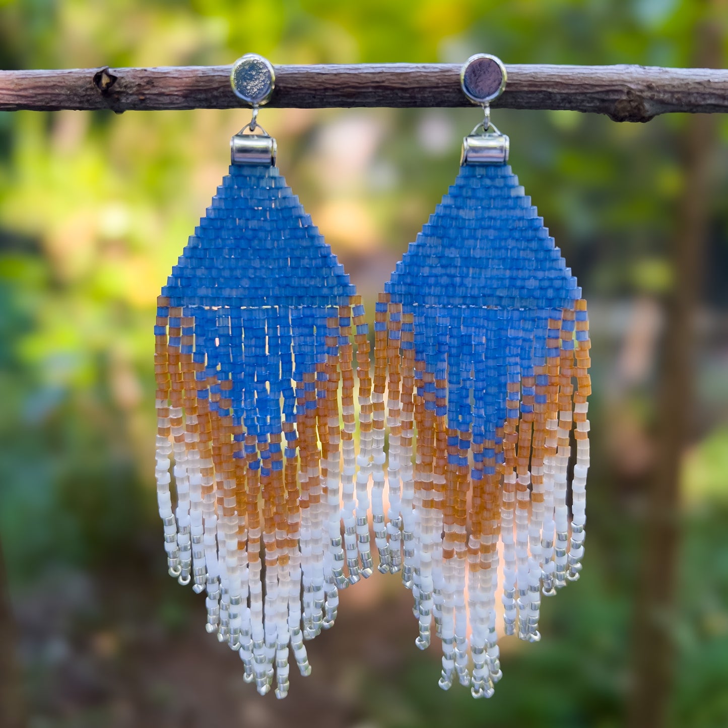 Pair of beaded Eastern Bluebird earrings hanging from a branch, featuring a vibrant blue triangular top with layered amber and white beaded fringe, creating a gradient effect from deep blue to warm amber and soft white.