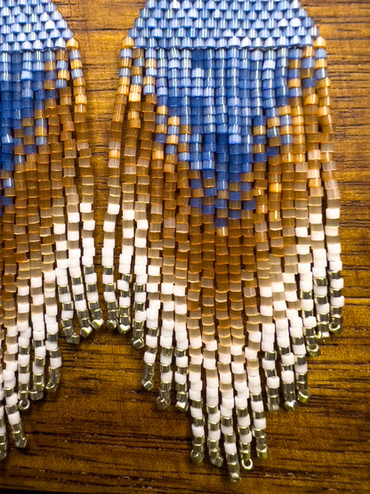 Close up of beaded Eastern Bluebird earrings on a wooden background, showcasing a blue triangular design at the top with cascading fringe in shades of amber and white, blending together in a striking ombré pattern.