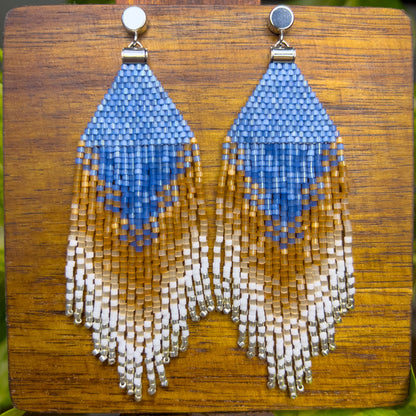 Beaded Eastern Bluebird earrings on a wooden background, showcasing a blue triangular design at the top with cascading fringe in shades of amber and white, blending together in a striking ombré pattern.