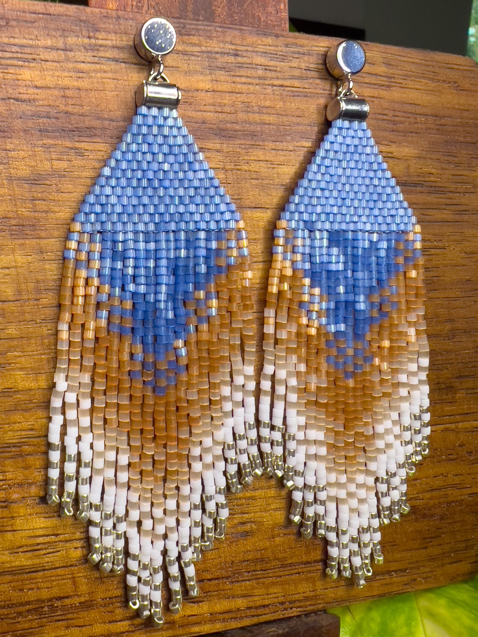 Side view of beaded Eastern Bluebird earrings on a wooden background, showcasing a blue triangular design at the top with cascading fringe in shades of amber and white, blending together in a striking ombré pattern.