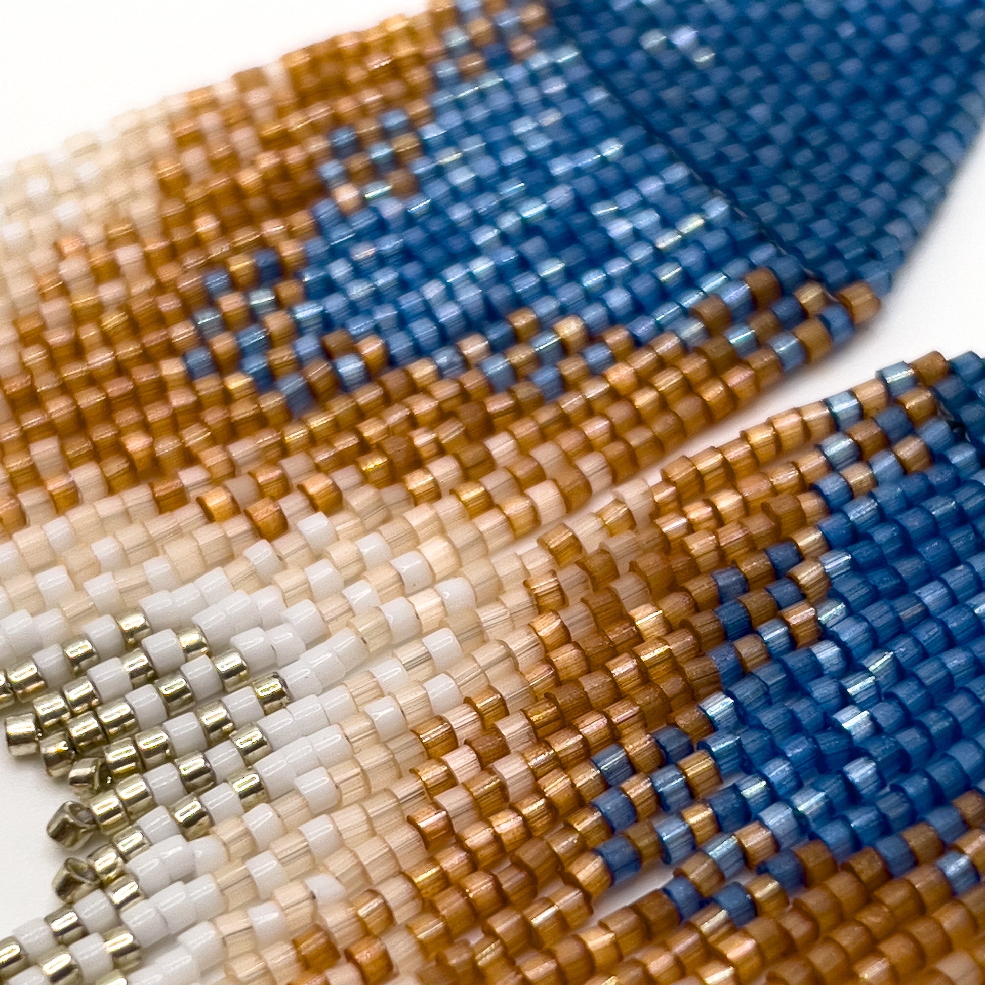 Close up of beaded Eastern Bluebird earrings on a white background, showcasing a blue triangular design at the top with cascading fringe in shades of amber and white, blending together in a striking ombré pattern.