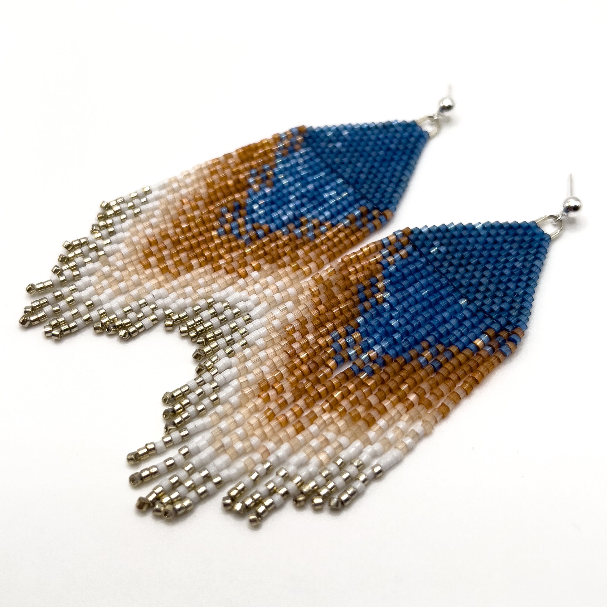 Close up of beaded Eastern Bluebird earrings on a white background, showcasing a blue triangular design at the top with cascading fringe in shades of amber and white, blending together in a striking ombré pattern.