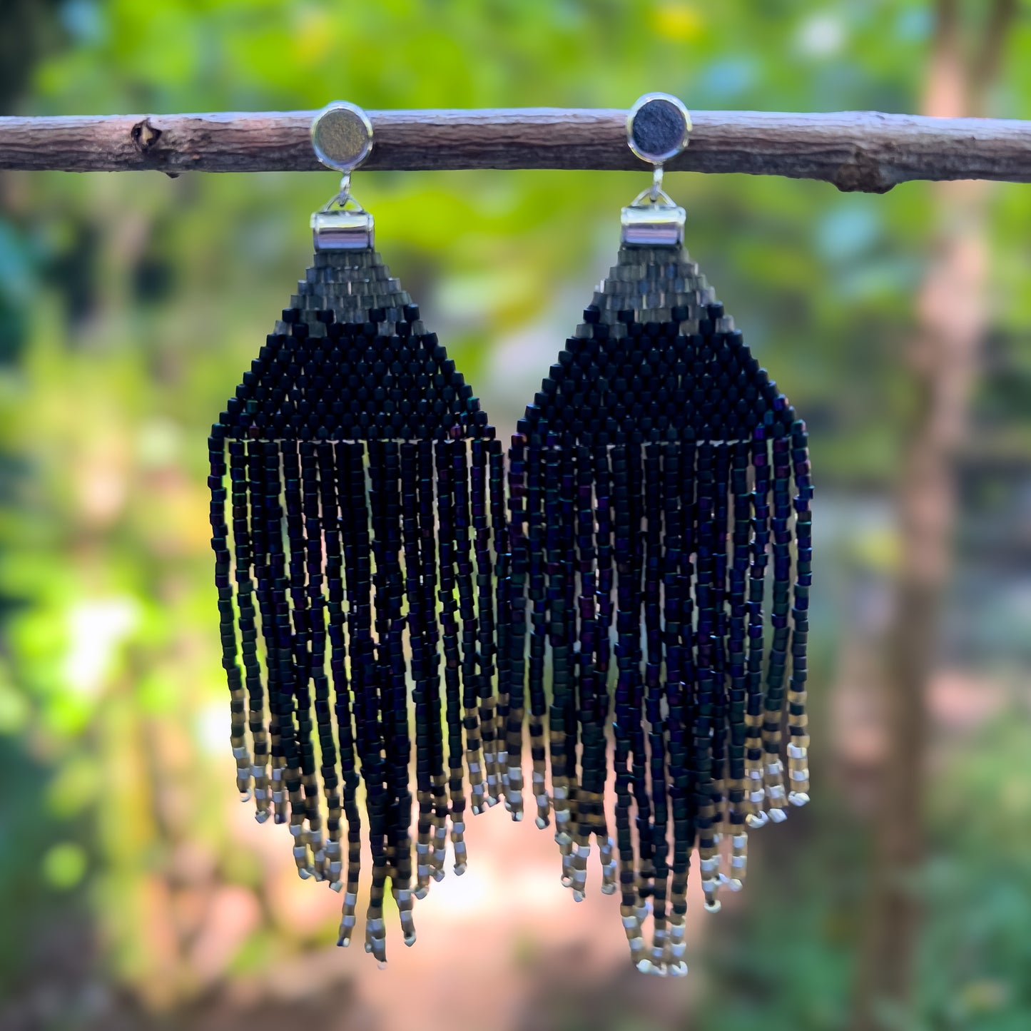 Pair of beaded earrings hanging from a branch, with a rich black and iridescent beaded top that transitions into long, layered fringe in dark, shimmering tones, capturing the colors and sheen of a European Starling.