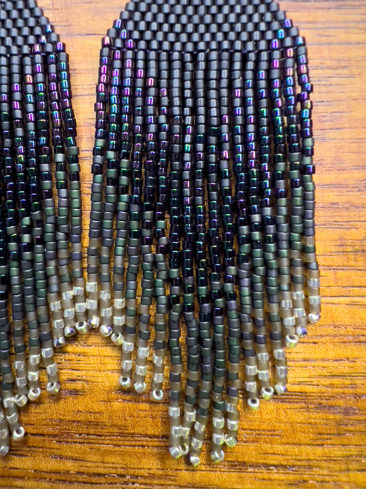 Close up of European Starling inspired earrings on a wooden background, showcasing a deep black beaded section that fades into iridescent fringe with hints of green and purple.