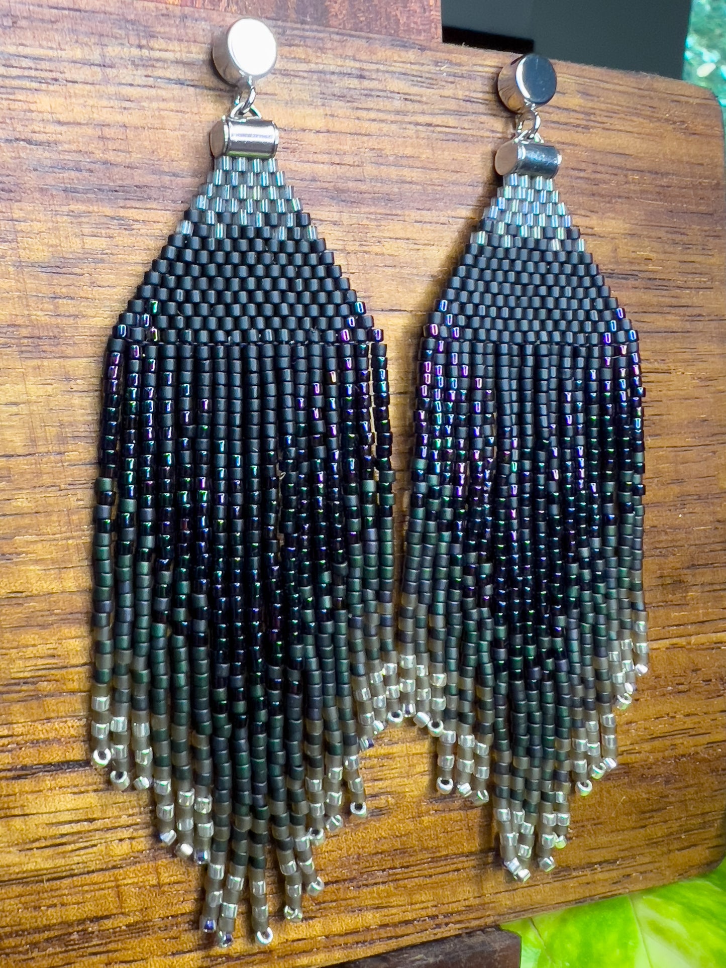 Side view of European Starling inspired earrings on a wooden background, showcasing a deep black beaded section that fades into iridescent fringe with hints of green and purple.