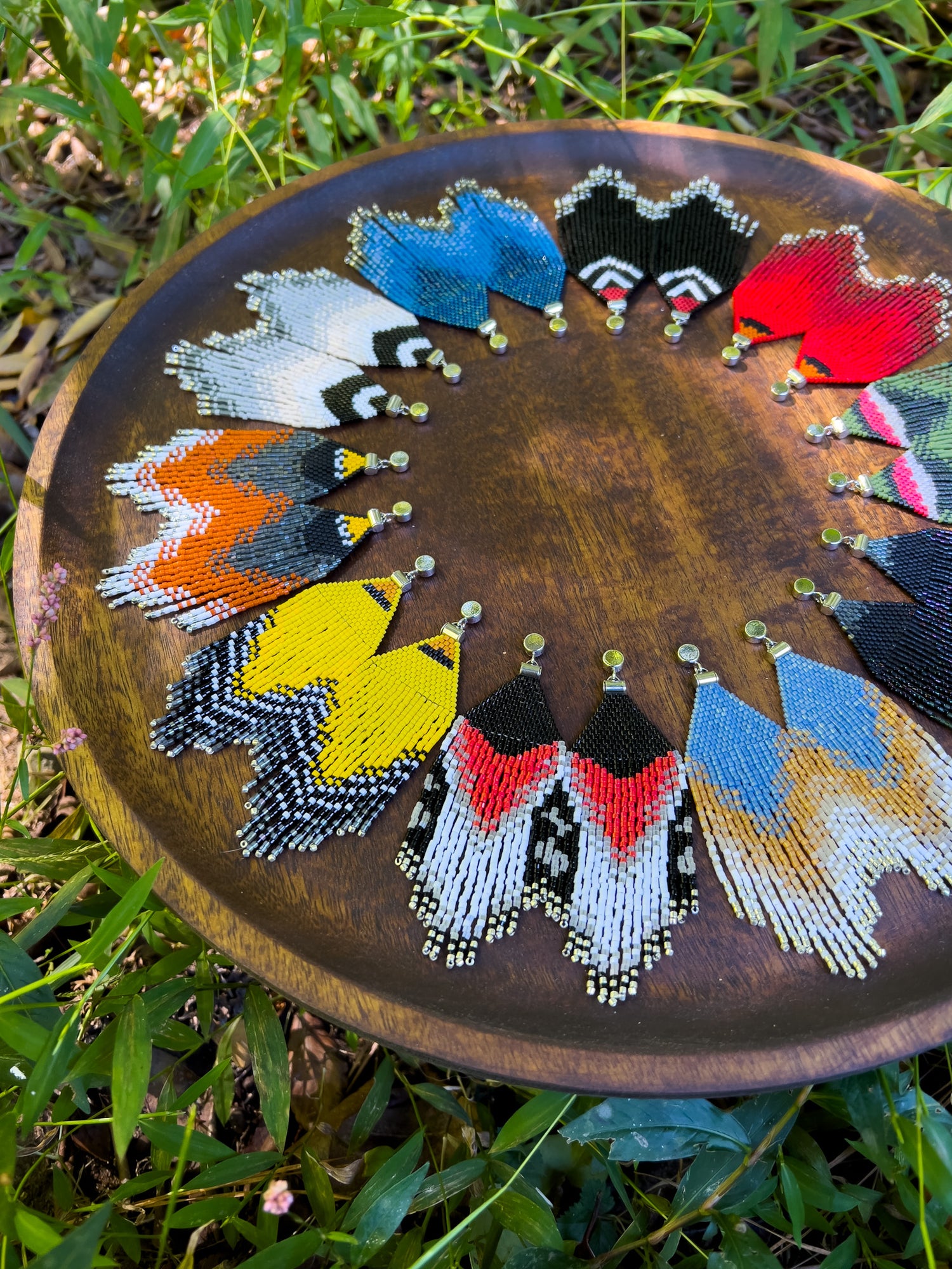 A close-up of a wooden tray with handcrafted beaded earrings arranged in a half-circle, featuring vibrant designs inspired by bird species, resting on a grassy background.