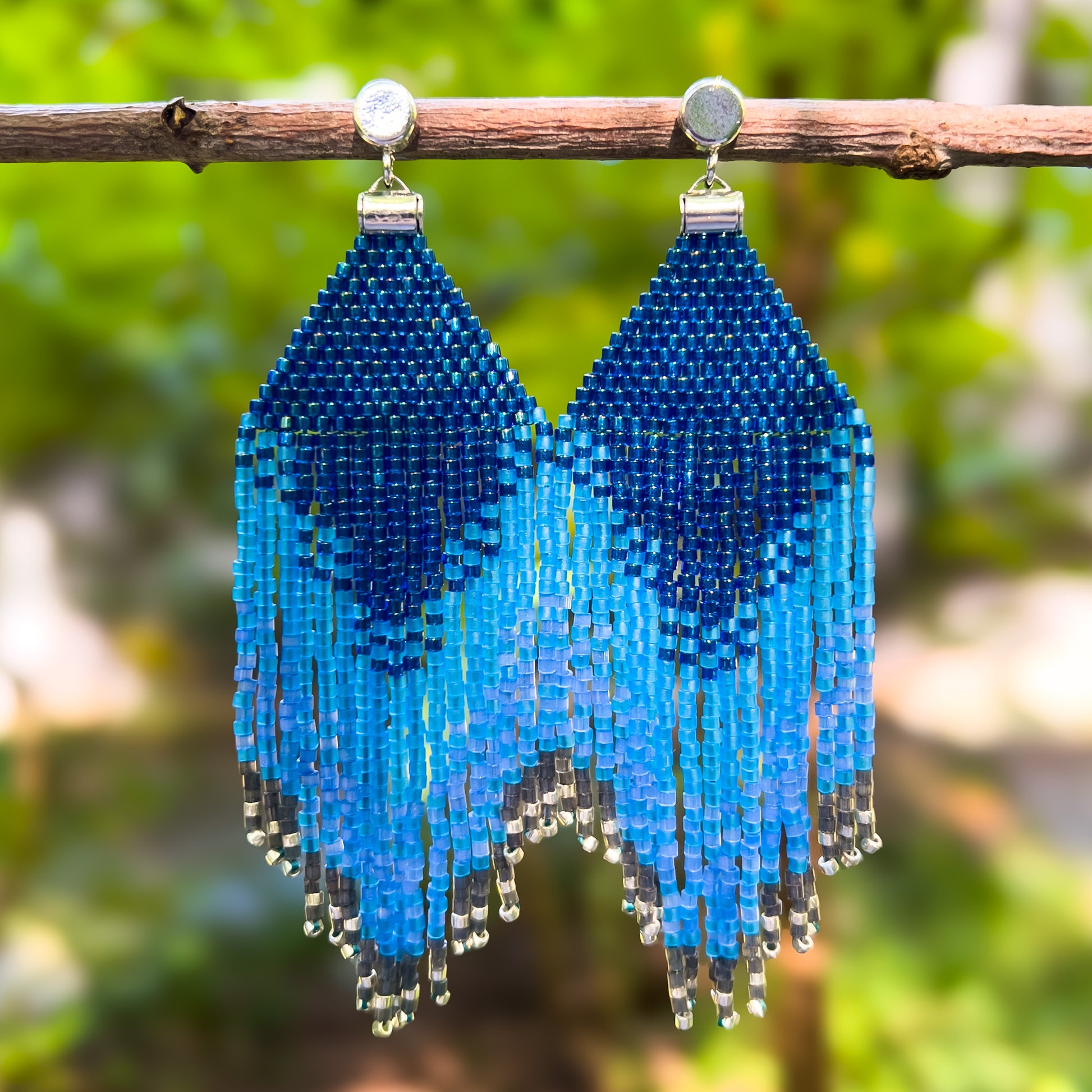 Pair of beaded Indigo Bunting earrings hanging from a branch, featuring a deep blue triangular top with cascading fringe in shades of bright and dark blue, creating a layered ombré effect that ends with silver bead accents.