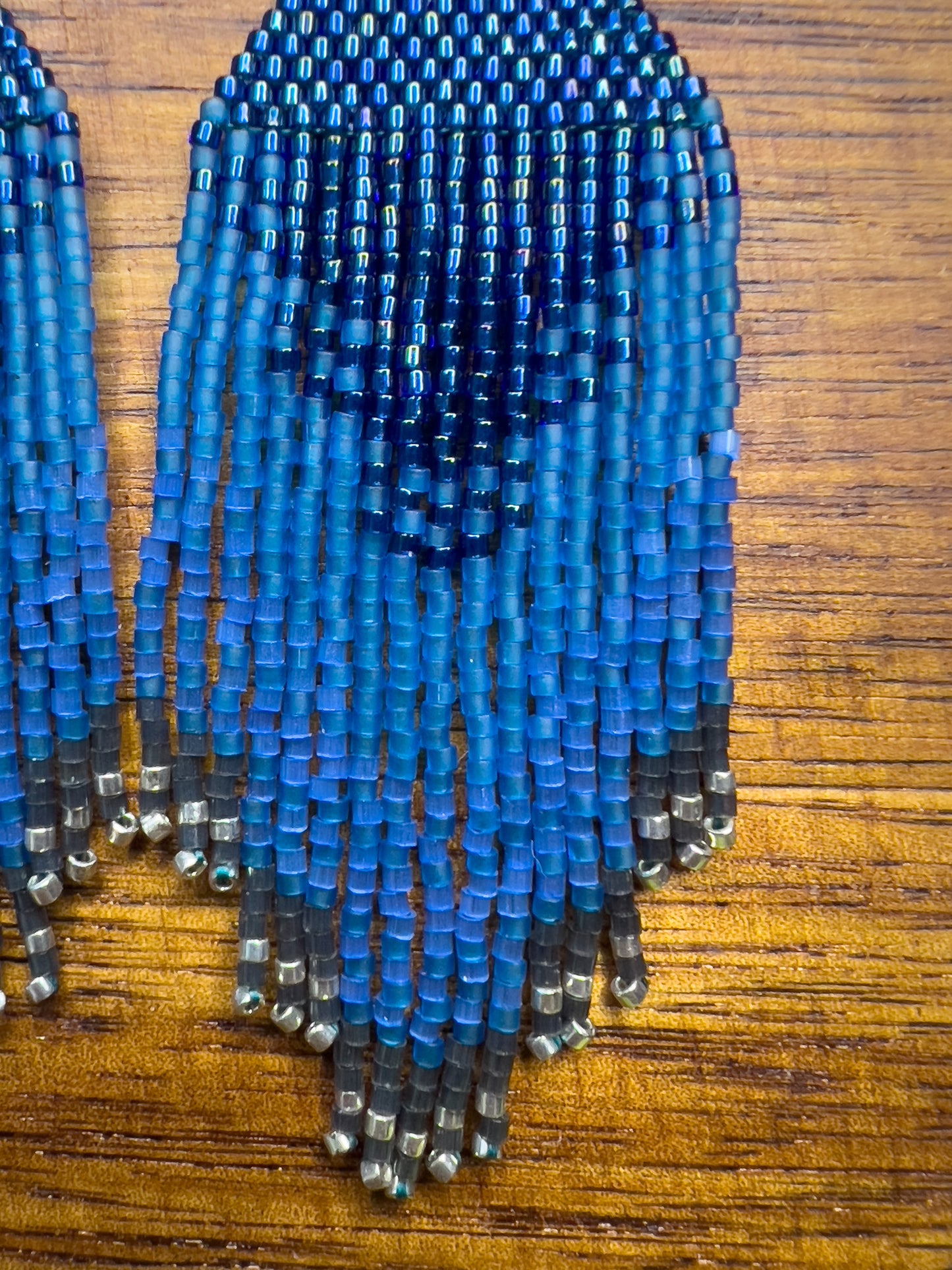 Close up of Indigo Bunting beaded earrings on a wooden background, showing a rich blue design at the top that transitions into long fringe with varying blue shades, tapering off with silver beads at the bottom.