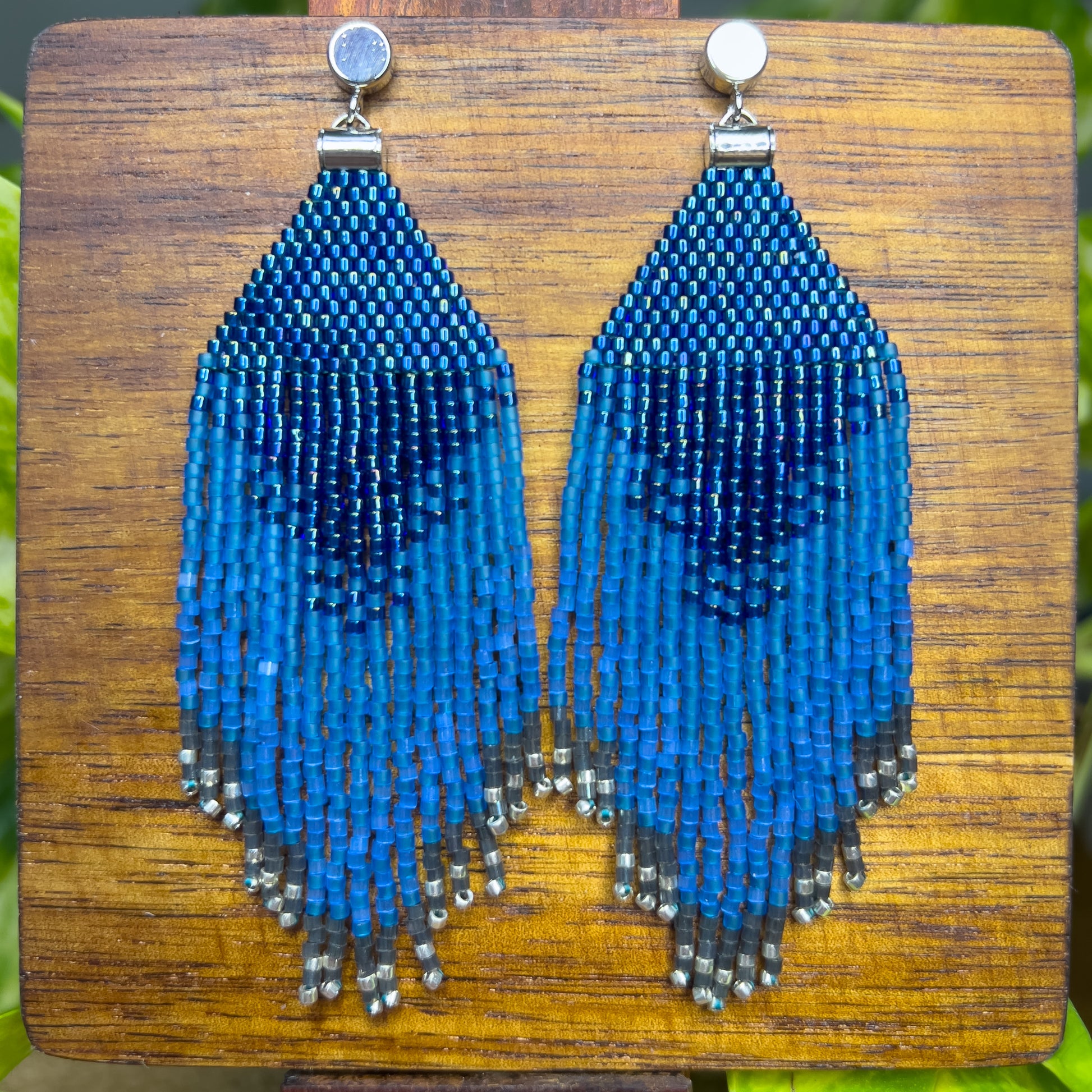 Indigo Bunting beaded earrings on a wooden background, showing a rich blue design at the top that transitions into long fringe with varying blue shades, tapering off with silver beads at the bottom.