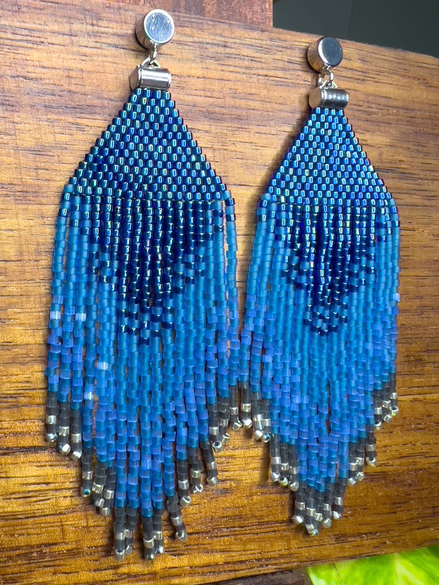 Side view of Indigo Bunting beaded earrings on a wooden background, showing a rich blue design at the top that transitions into long fringe with varying blue shades, tapering off with silver beads at the bottom.