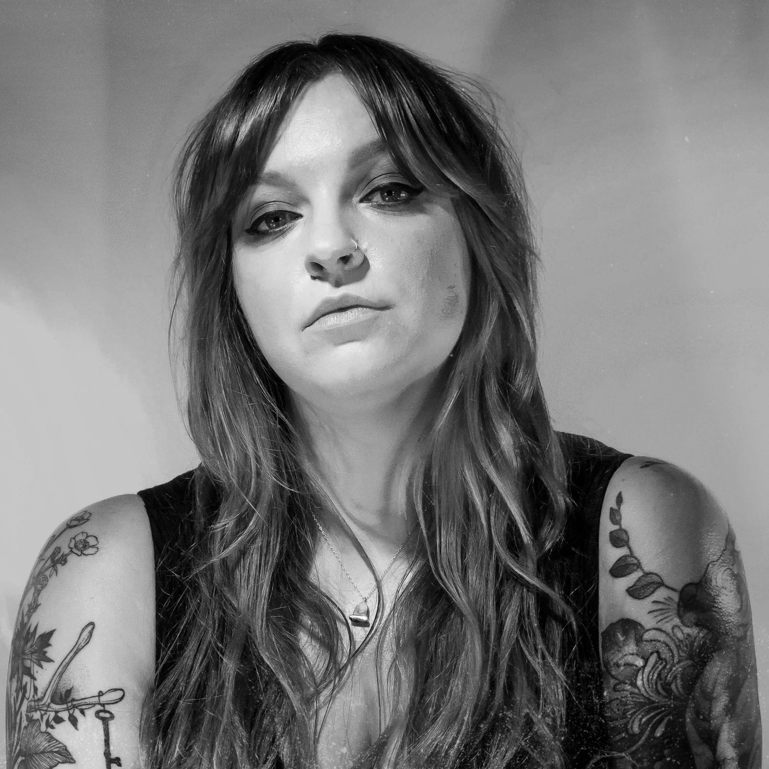 Black and white portrait of a woman with long wavy hair and tattoos on both arms, gazing directly at the camera with a calm expression.