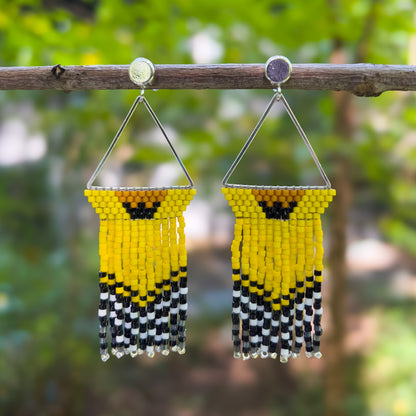 Handcrafted beaded earrings inspired by the American Goldfinch, featuring a triangular design with yellow, black, and white beads, hanging from a branch against a blurred natural background.