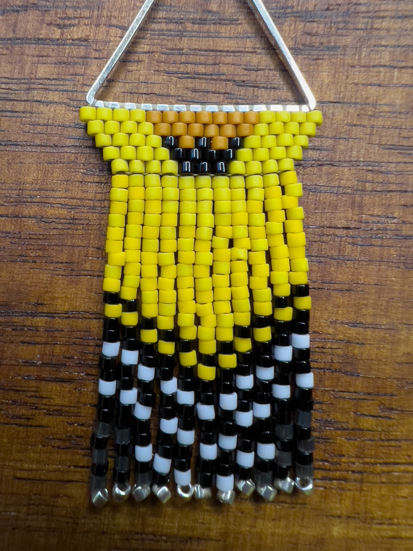 Close up of American Goldfinch-inspired handcrafted beaded earrings displayed on a wooden surface, showcasing a triangular design with bright yellow, black, and white beaded patterns and fringe.