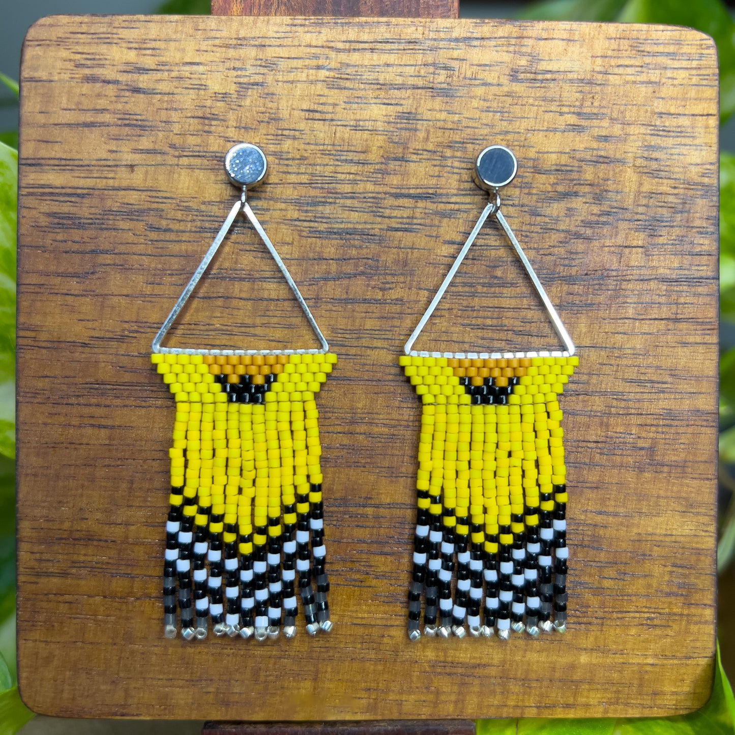 American Goldfinch-inspired handcrafted beaded earrings displayed on a wooden surface, showcasing a triangular design with bright yellow, black, and white beaded patterns and fringe.