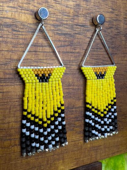 Side view of American Goldfinch-inspired handcrafted beaded earrings displayed on a wooden surface, showcasing a triangular design with bright yellow, black, and white beaded patterns and fringe.