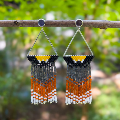 Handcrafted beaded earrings inspired by the American Robin featuring a triangular design with shades of yellow, black, orange, and white beads, hanging from a branch against a blurred natural background.