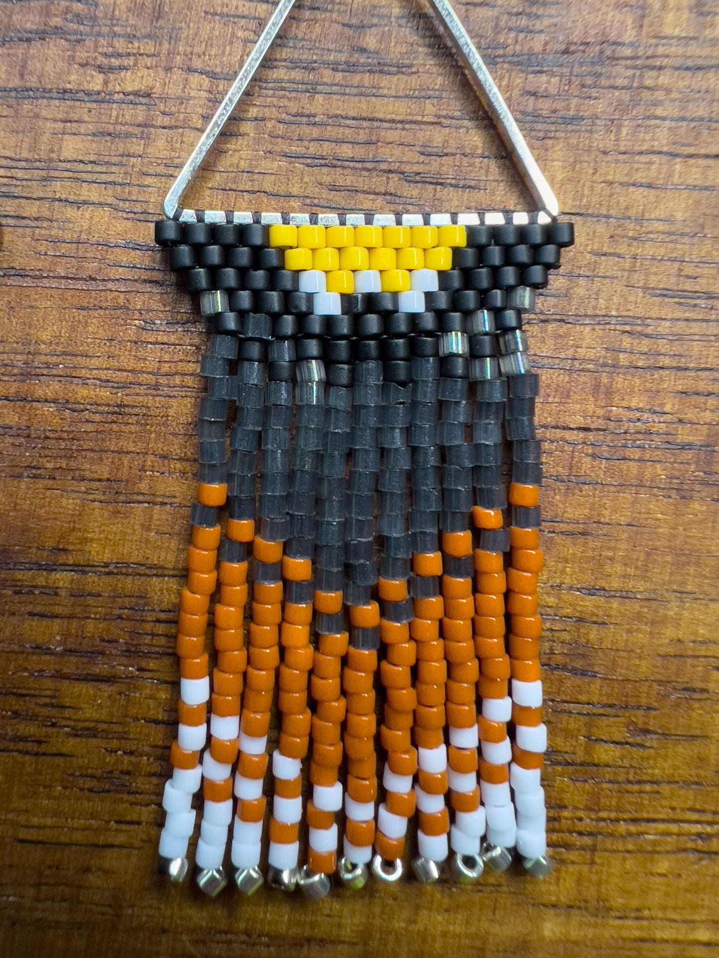 Close up of handcrafted beaded earrings inspired by an American Robin displayed on a wooden surface, showcasing a triangular design with yellow, black, orange, and white beads in a vibrant, nature-inspired pattern.