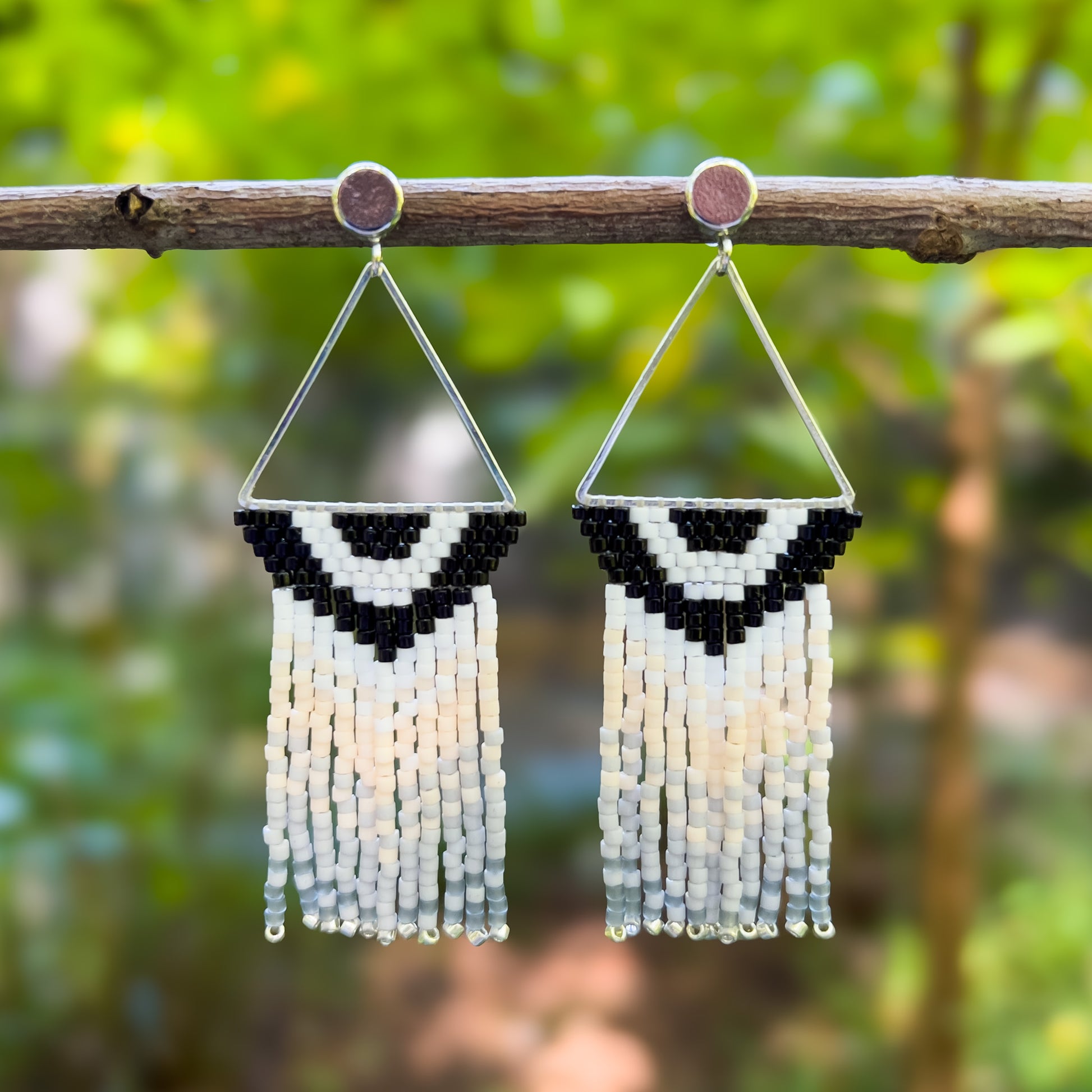 Pair of beaded earrings inspired by the Carolina Chickadee, hanging from a branch, featuring a triangular silver frame with a black and white V-shaped bead pattern at the top and cascading fringe in shades of cream and gray.