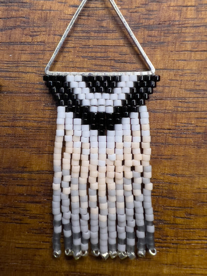 Close up of Carolina Chickadee inspired earrings on a wooden background, showing a triangular silver frame holding a black and white V-patterned beadwork with long cream and gray beaded fringe hanging below.