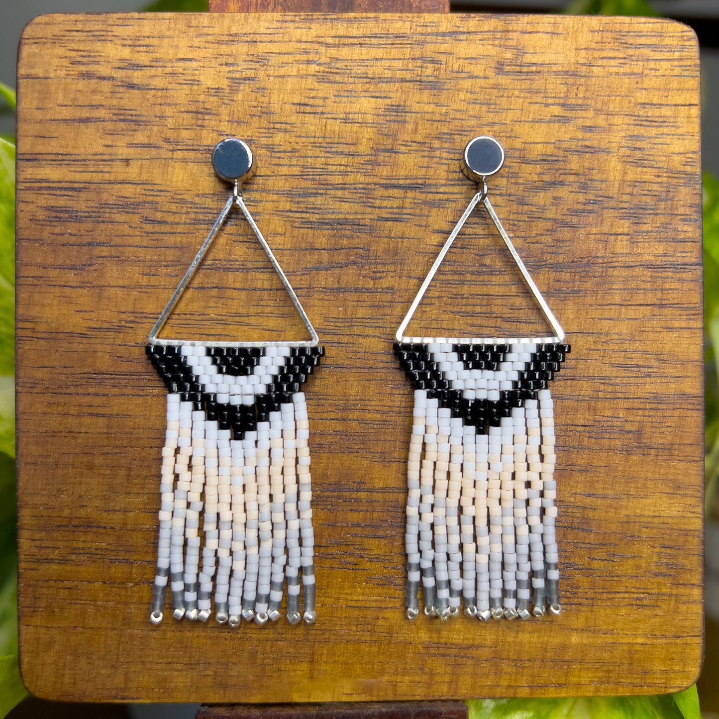 Carolina Chickadee-inspired earrings on a wooden background, showing a triangular silver frame holding a black and white V-patterned beadwork with long cream and gray beaded fringe hanging below.