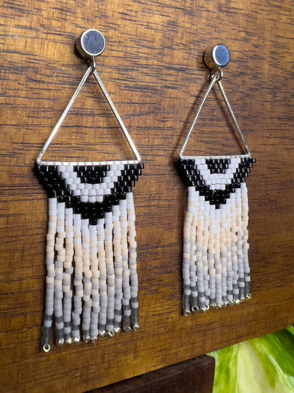 Side view of Carolina Chickadee inspired earrings on a wooden background, showing a triangular silver frame holding a black and white V-patterned beadwork with long cream and gray beaded fringe hanging below.