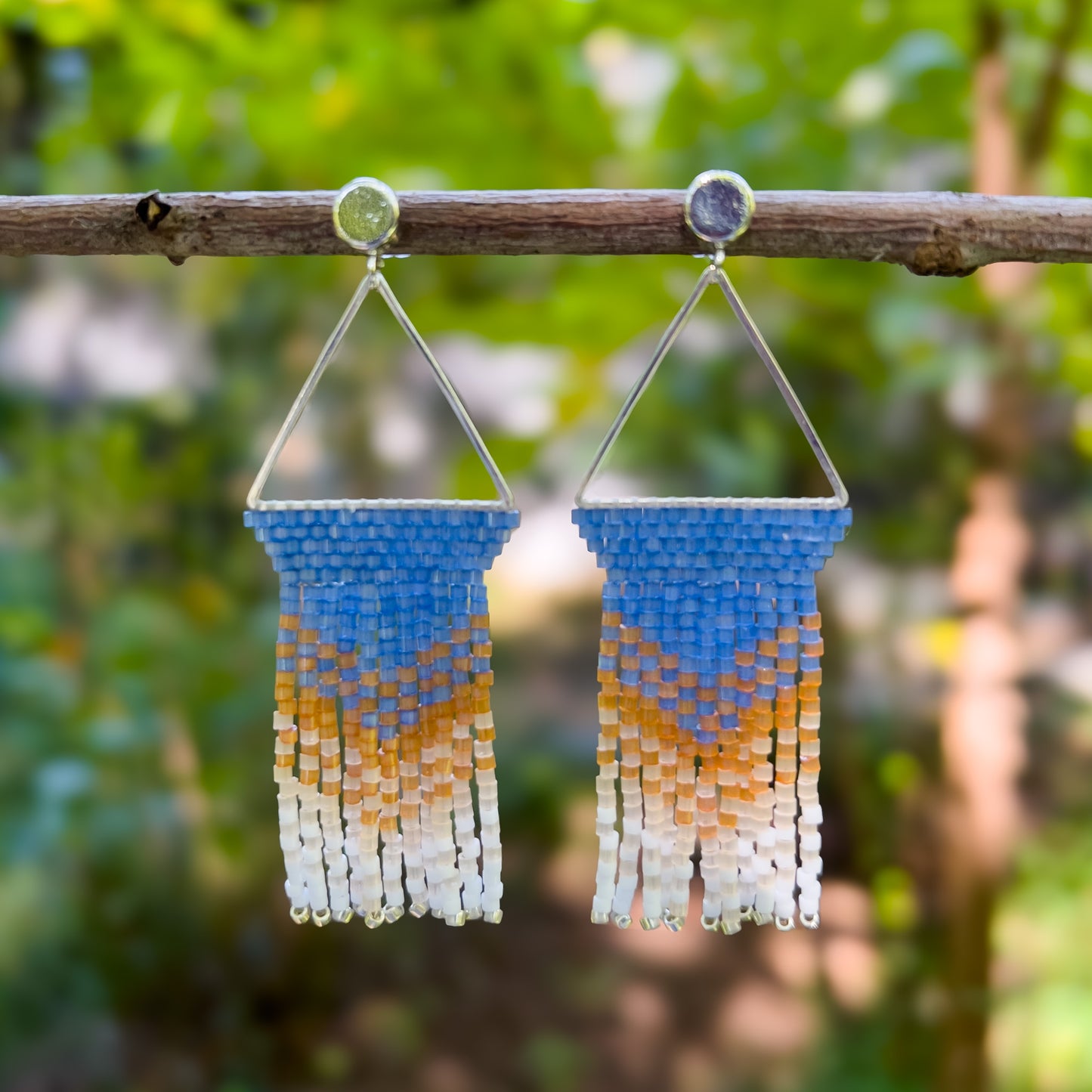 Pair of beaded Eastern Bluebird earrings hanging from a branch, featuring a triangular silver frame with a solid blue beaded top that transitions into amber and white beaded fringe, creating a gentle gradient effect.