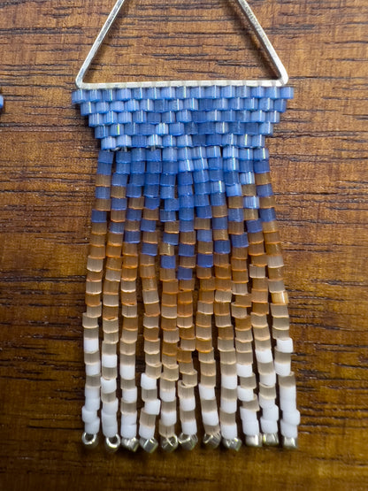 Close up of beaded Eastern Bluebird earrings on a wooden background, showcasing a blue beaded section within a triangular silver frame, with cascading fringe in amber and white for a layered, ombré look.