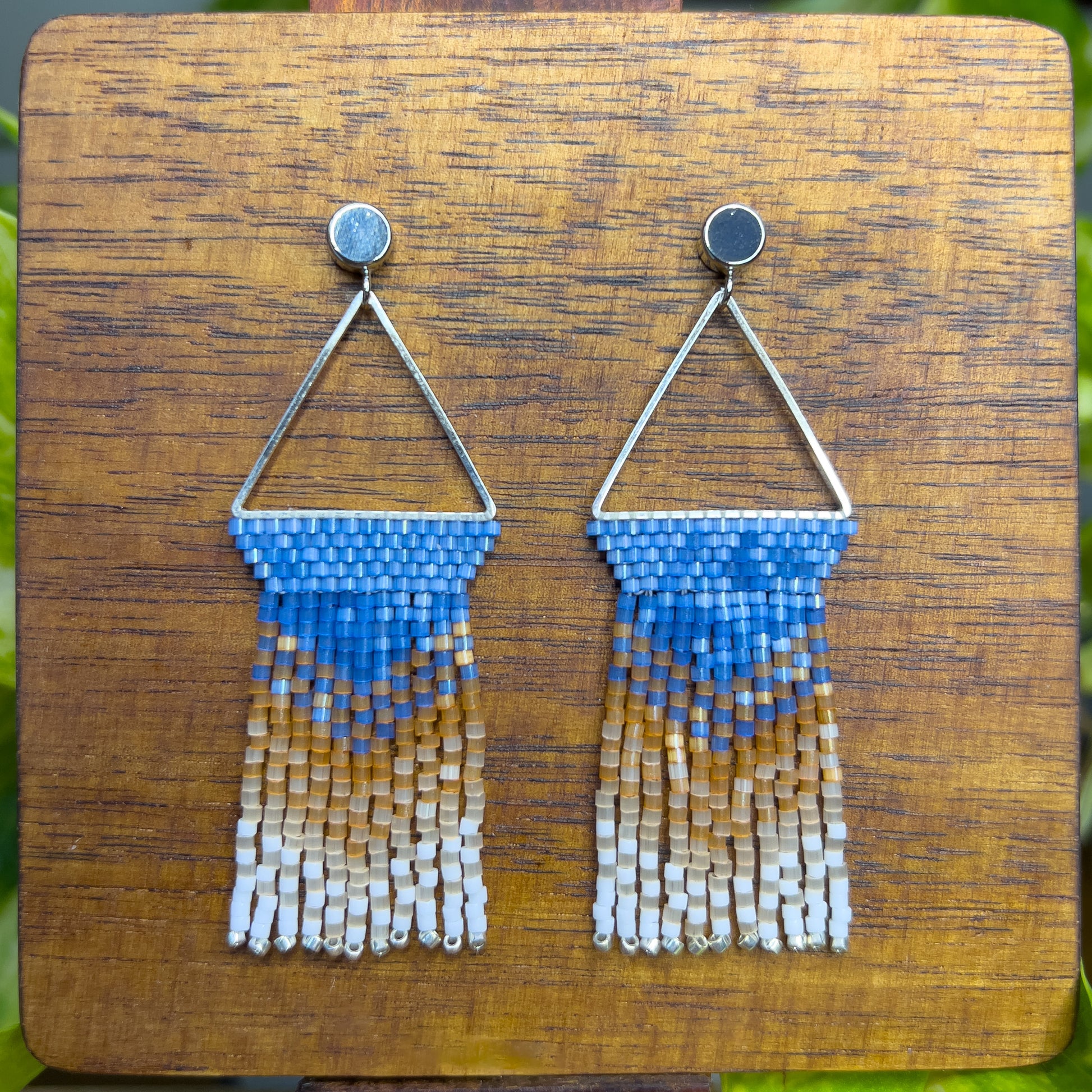 Beaded Eastern Bluebird earrings on a wooden background, showcasing a blue beaded section within a triangular silver frame, with cascading fringe in amber and white for a layered, ombré look.