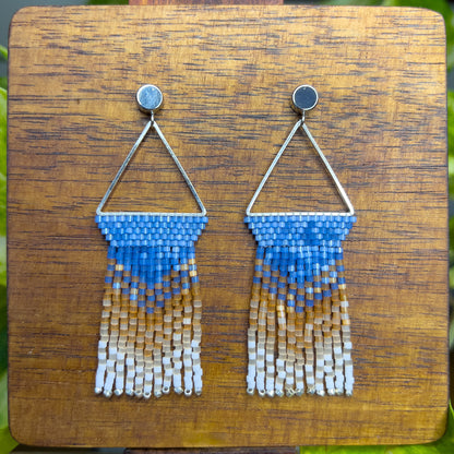 Beaded Eastern Bluebird earrings on a wooden background, showcasing a blue beaded section within a triangular silver frame, with cascading fringe in amber and white for a layered, ombré look.