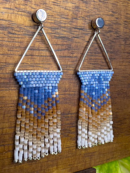 Side view of beaded Eastern Bluebird earrings on a wooden background, showcasing a blue beaded section within a triangular silver frame, with cascading fringe in amber and white for a layered, ombré look.