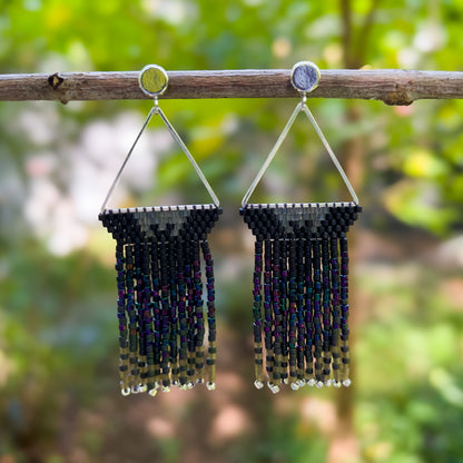 Pair of beaded earrings hanging from a branch, with a triangular silver frame holding black beaded sections that transition into dark, iridescent fringe featuring shades of green and purple, capturing the shimmering colors of a European Starling.