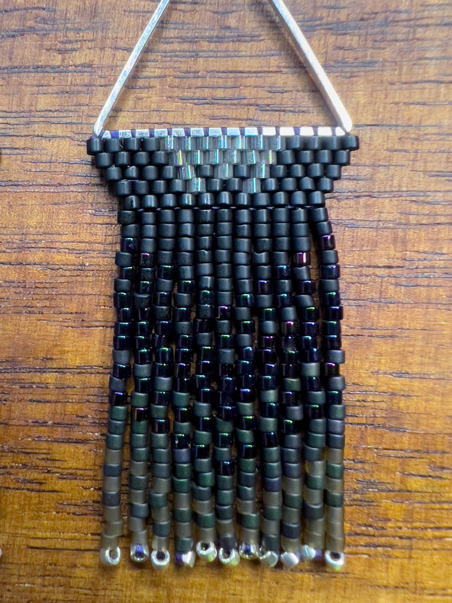 Close up of European Starling inspired earrings on a wooden background, showcasing black beaded tops within a triangular silver frame, with long, iridescent fringe that shifts from black to hints of green and purple.