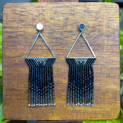 European Starling inspired earrings on a wooden background, showcasing black beaded tops within a triangular silver frame, with long, iridescent fringe that shifts from black to hints of green and purple.