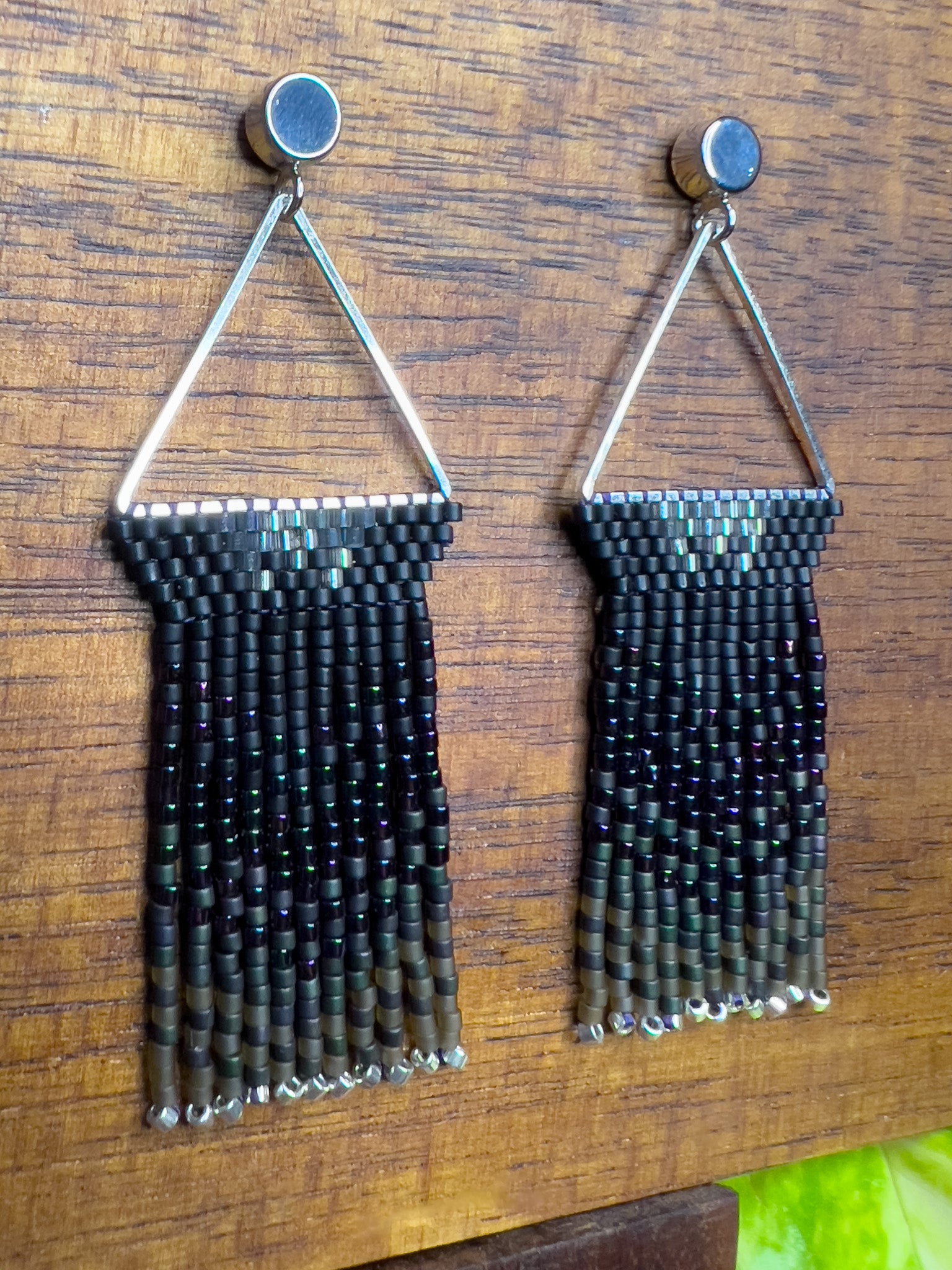 Side view of European Starling inspired earrings on a wooden background, showcasing black beaded tops within a triangular silver frame, with long, iridescent fringe that shifts from black to hints of green and purple.