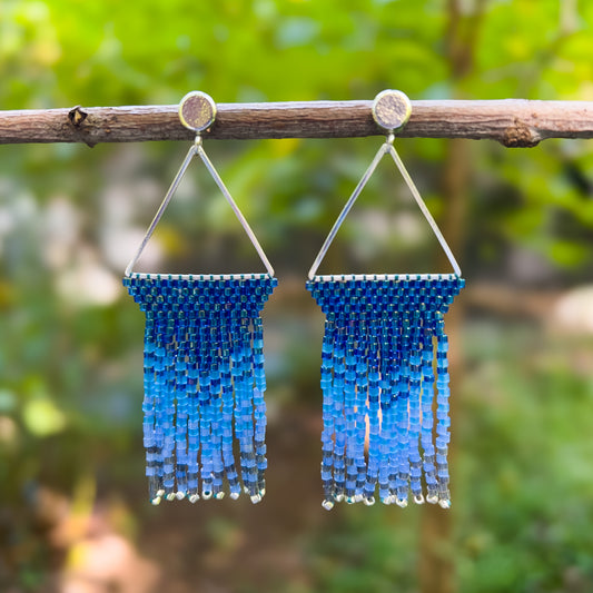 Pair of Indigo Bunting beaded earrings hanging from a branch, featuring a triangular silver frame with tightly woven blue beads at the top, cascading into a gradient of deep and light blue fringe with silver bead accents.