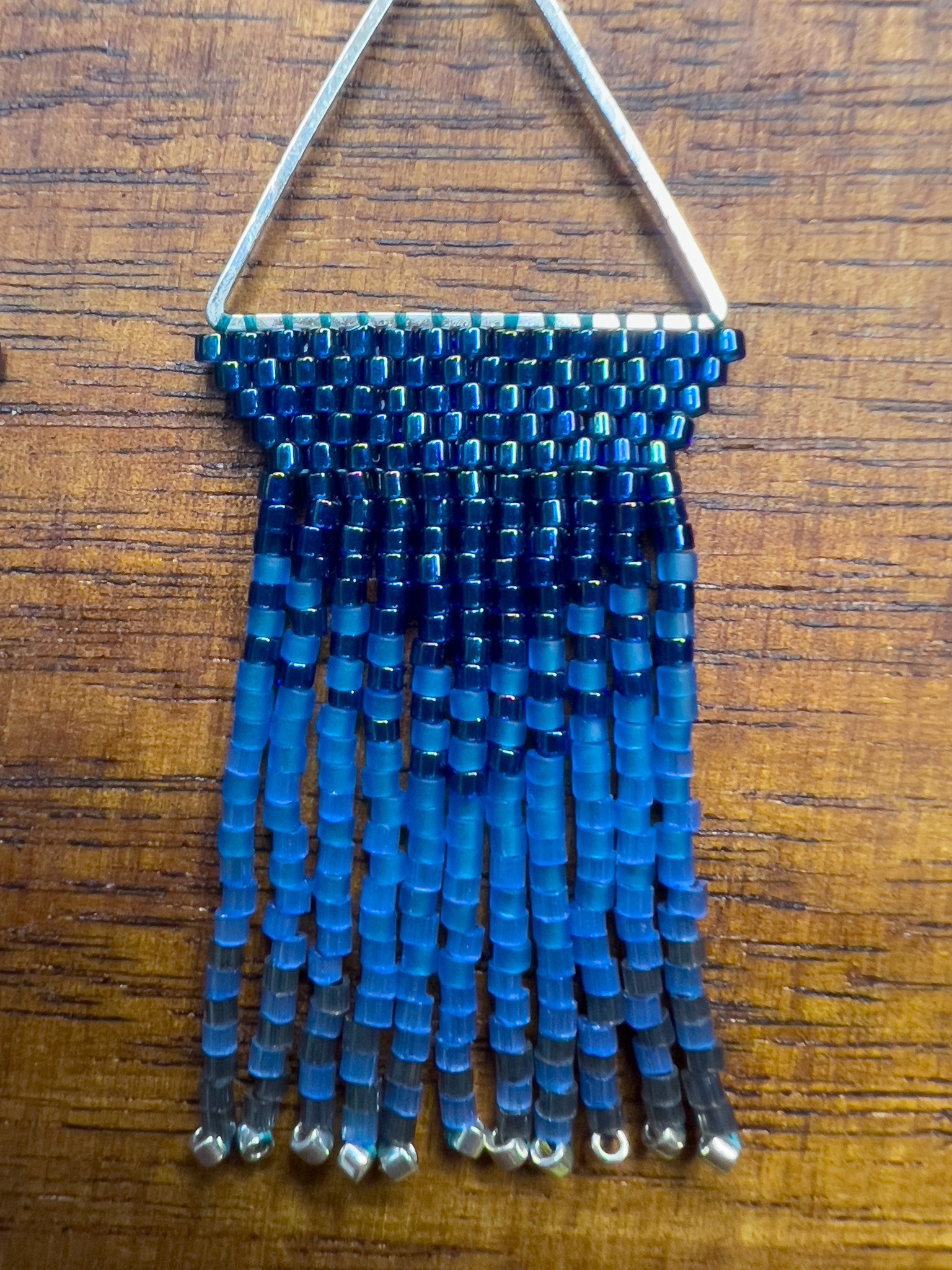 Close up of Indigo Bunting earrings on a wooden background, showcasing a silver triangular frame holding rows of blue beads that transition into lighter blue fringe with silver bead accents at the ends.