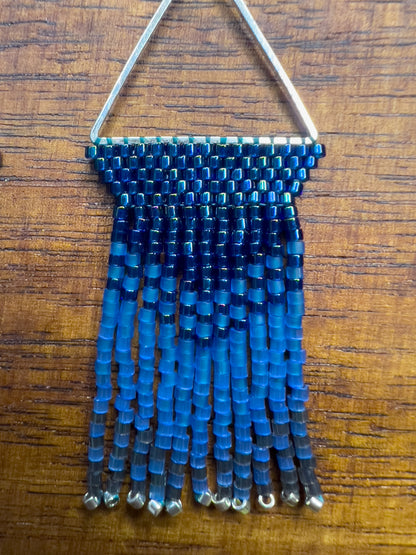 Close up of Indigo Bunting earrings on a wooden background, showcasing a silver triangular frame holding rows of blue beads that transition into lighter blue fringe with silver bead accents at the ends.