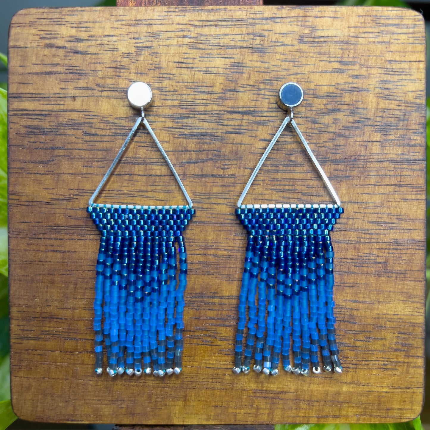 Beaded Indigo Bunting earrings on a wooden background, showcasing a silver triangular frame holding rows of blue beads that transition into lighter blue fringe with silver bead accents at the ends.