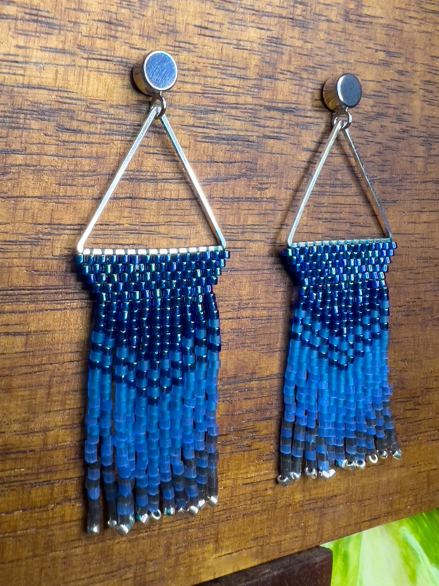 Side view of Indigo Bunting earrings on a wooden background, showcasing a silver triangular frame holding rows of blue beads that transition into lighter blue fringe with silver bead accents at the ends.