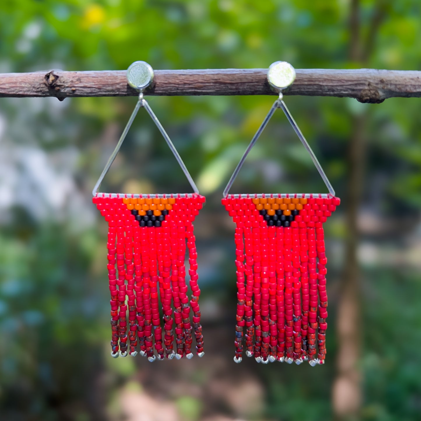 Pair of red Northern Cardinal beaded earrings hanging from a branch, featuring a triangular silver frame with a red beaded top accented by an orange and black section resembling a cardinal’s face, followed by a cascade of red fringe that ends in silver bead tips.